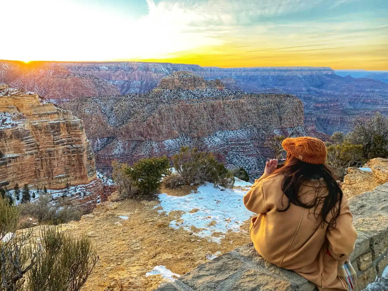 Woman in brown coat sits on cliff edge, gazing at Grand Canyon's vast beauty.